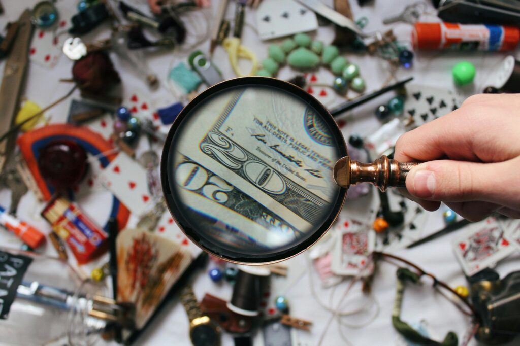 A hand holding a magnifying glass focusing on a twenty dollar bill amidst various scattered objects.